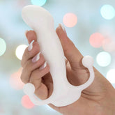 A hand with manicured nails holds the Aneros Progasm Prostate Massager - White, featuring its signature two rounded handles at the base. The background is blurred with colorful lights, creating a festive atmosphere.